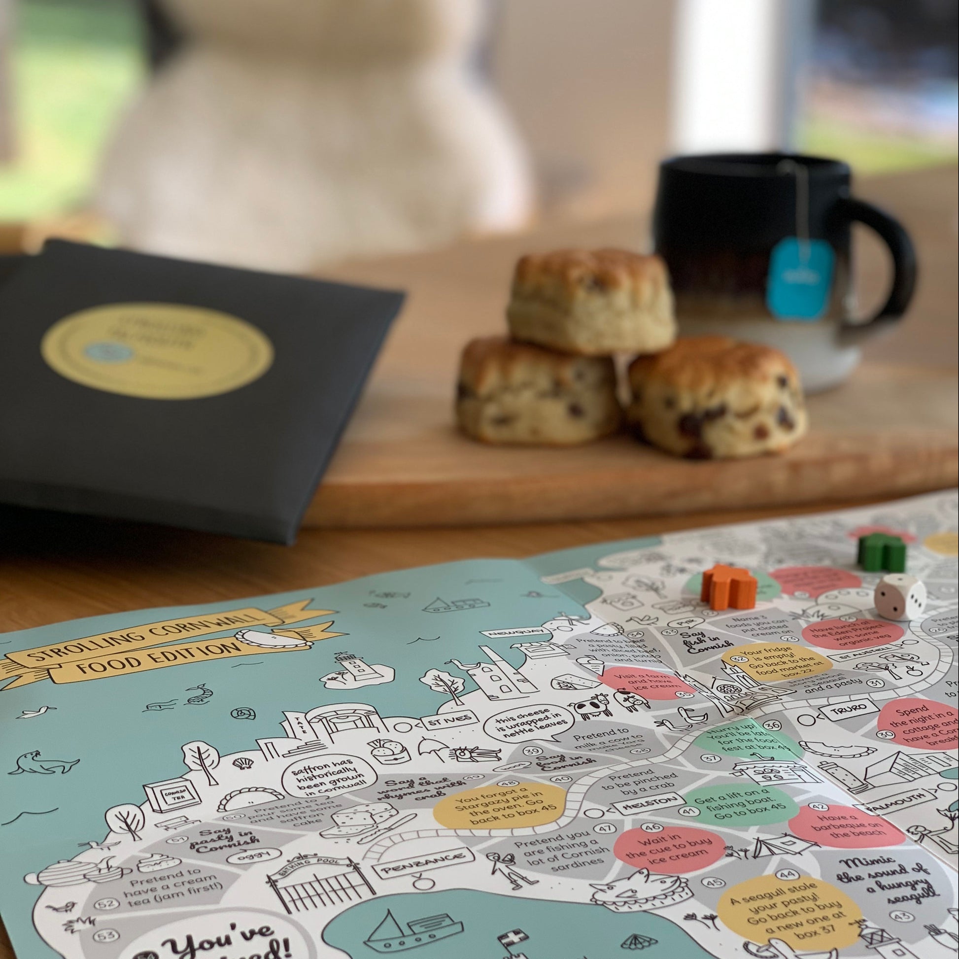 Cornwall board game food edition open on the table with wooden dice and markers. In the background some scones and Cornish cream tea.